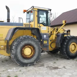 Żółta ładowarka kołowa Komatsu WA470 z widocznymi śladami użytkowania, stojąca na betonowym placu przed budynkiem z brązowym dachem.