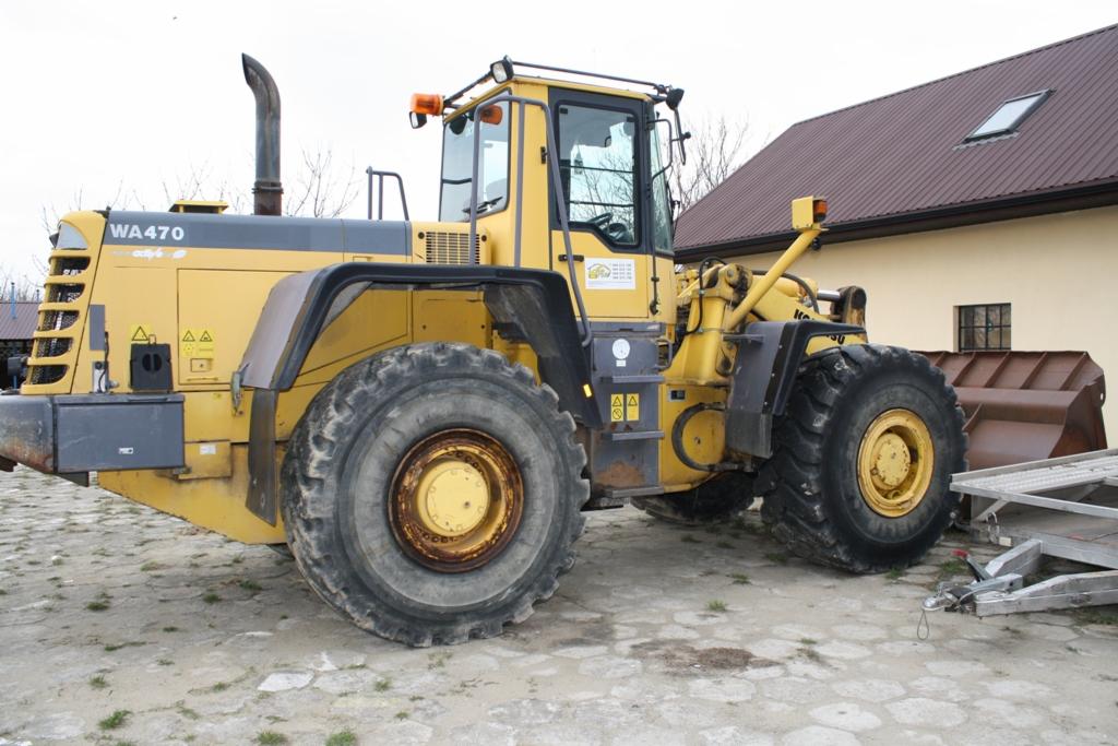 Żółta ładowarka kołowa Komatsu WA470 z widocznymi śladami użytkowania, stojąca na betonowym placu przed budynkiem z brązowym dachem.