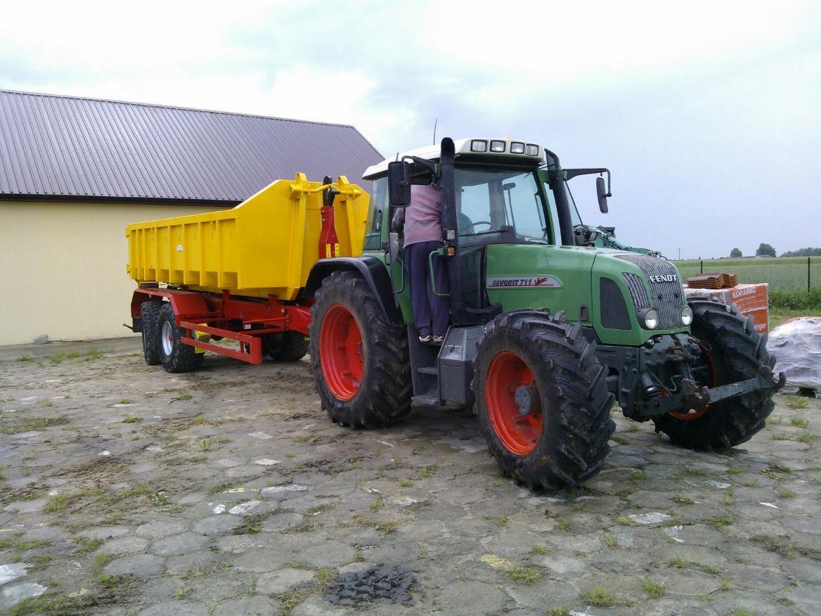 Zielony ciągnik Fendt Favorit 711 z żółtą przyczepą kontenerową na czerwonym podwoziu, stojący na betonowym placu przed budynkiem.