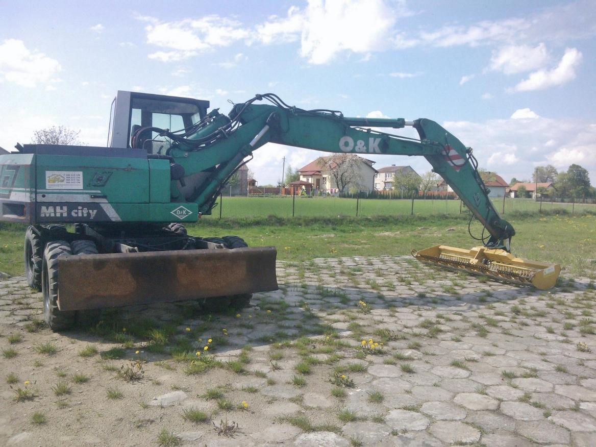 Zielona koparka kołowa O&K MH City z przodu z zamontowanym osprzętem do koszenia trawy, stojąca na betonowej nawierzchni, w tle trawnik i zabudowania.