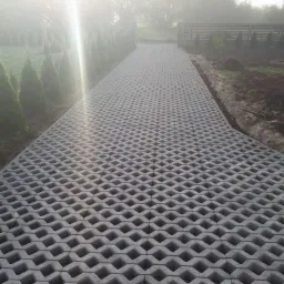 Długa, szara droga z ażurowych betonowych płyt prowadząca przez ogród w porannej mgle, obok rosną tuje i krzewy.