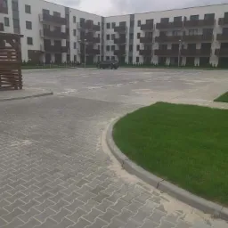 Nowo ułożona szara kostka brukowa na parkingu przed nowoczesnym budynkiem mieszkalnym z brązowymi balkonami; widoczne fragmenty trawnika i zaparkowany samochód w tle.