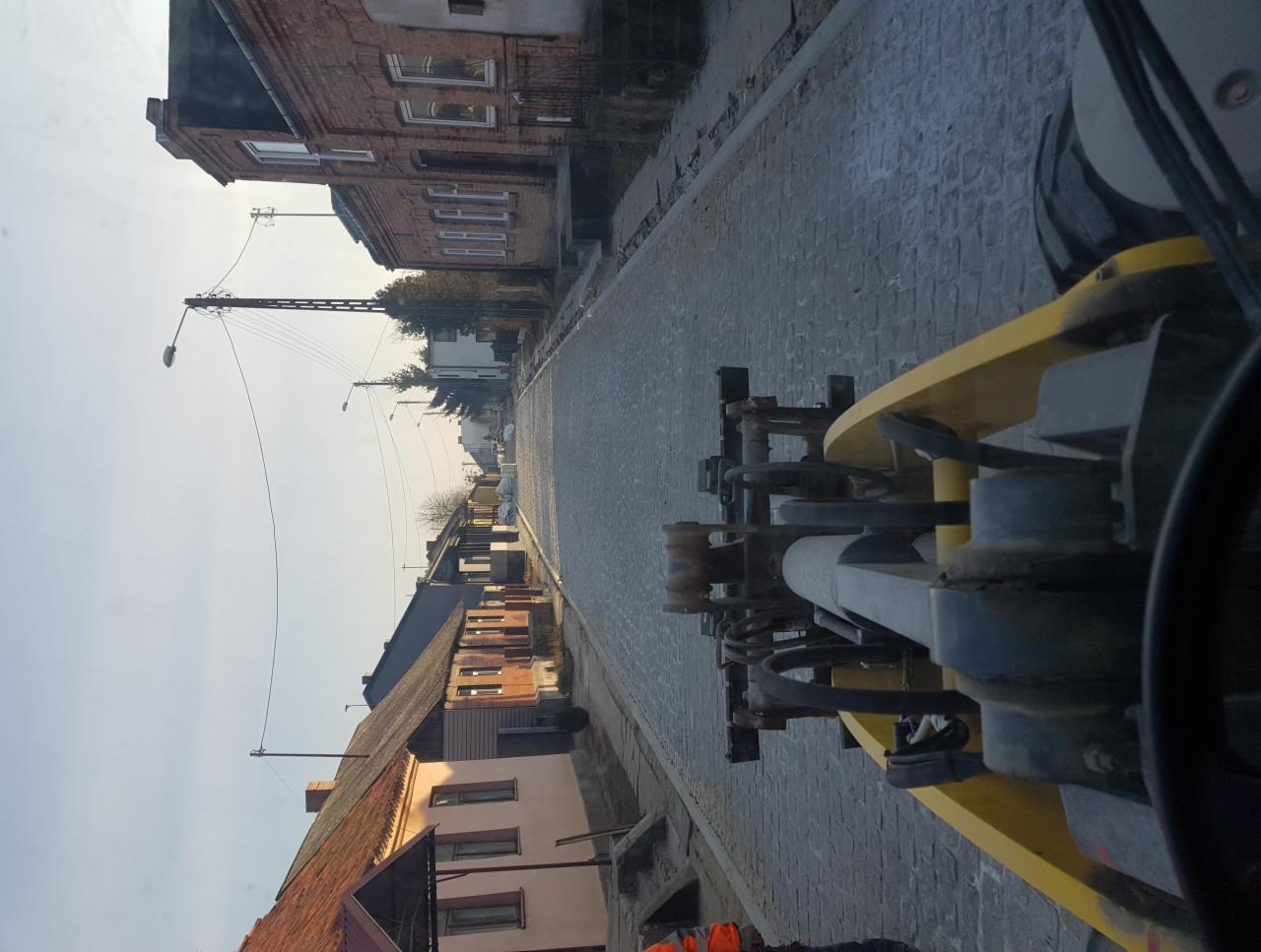 Widok z kabiny walca drogowego na świeżo ułożoną nawierzchnię z kostki brukowej na tle zabudowań miejskich w pochmurny dzień.