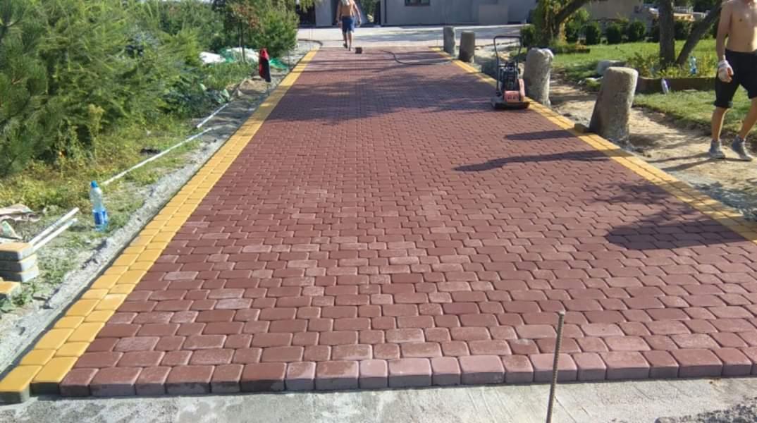 Czerwona kostka brukowa ułożona wzdłuż ścieżki z żółtym obramowaniem, widoczny zagęszczarka, mężczyzna w oddali i niedokończone krawędzie.