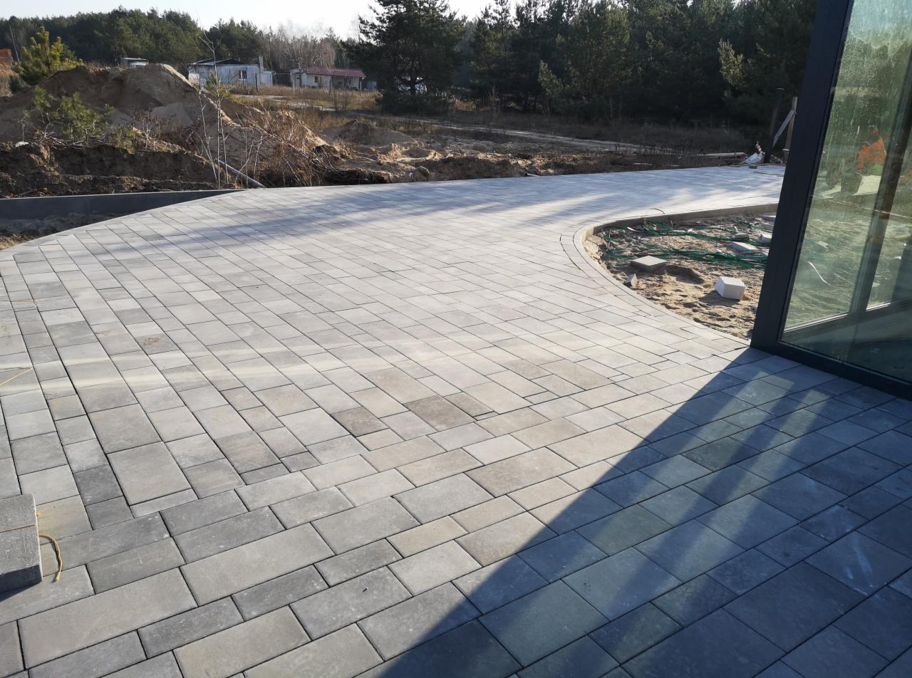Zakrzywiony podjazd z szarej kostki brukowej w kształcie prostokątów, częściowo zacieniony, z widocznym terenem budowy w tle i oknem budynku po prawej stronie.