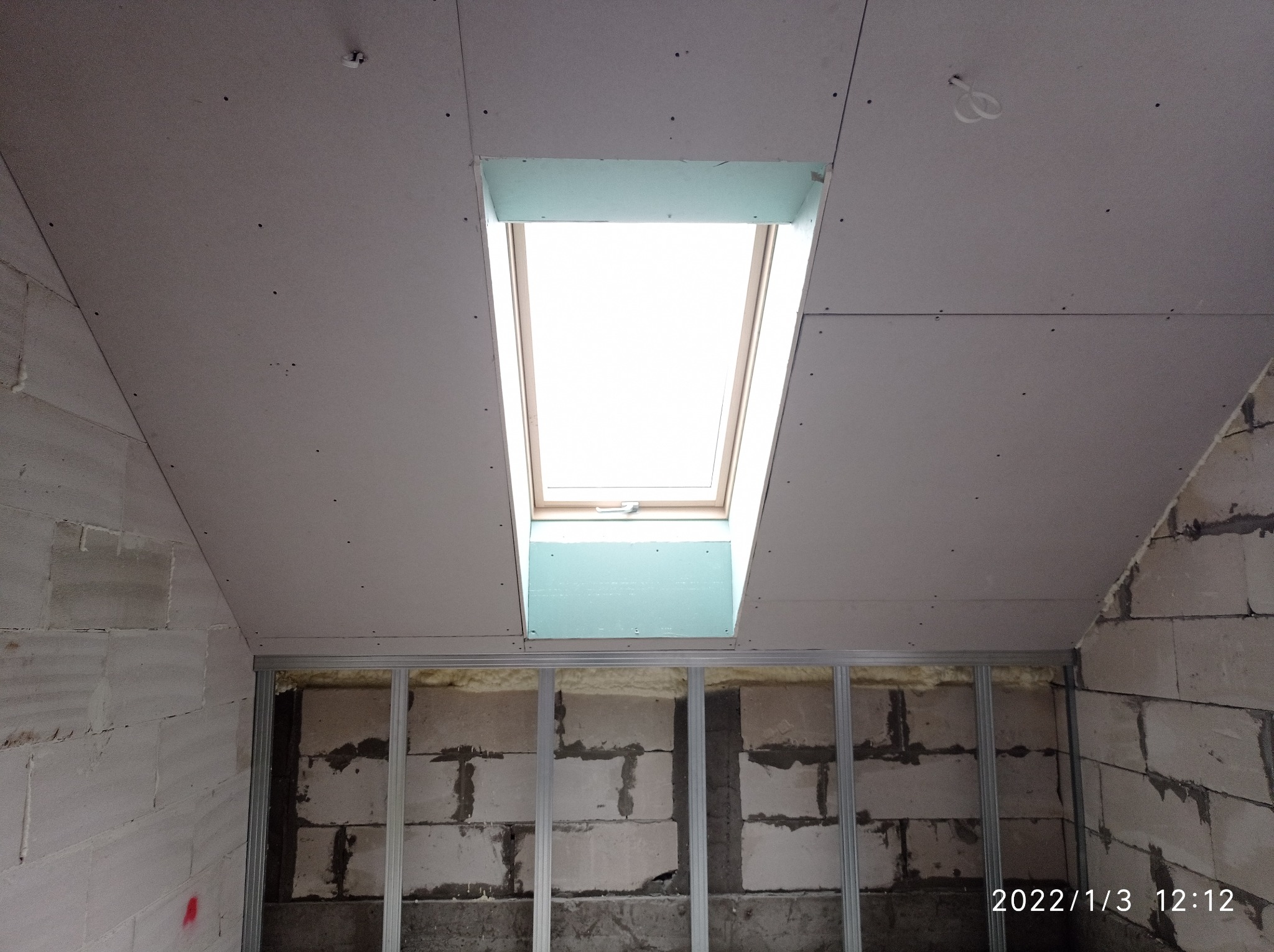 Montaż płyty kartonowo-gipsowej na suficie poddasza z oknem dachowym; widoczne profile metalowe i surowe ściany z betonu komórkowego.