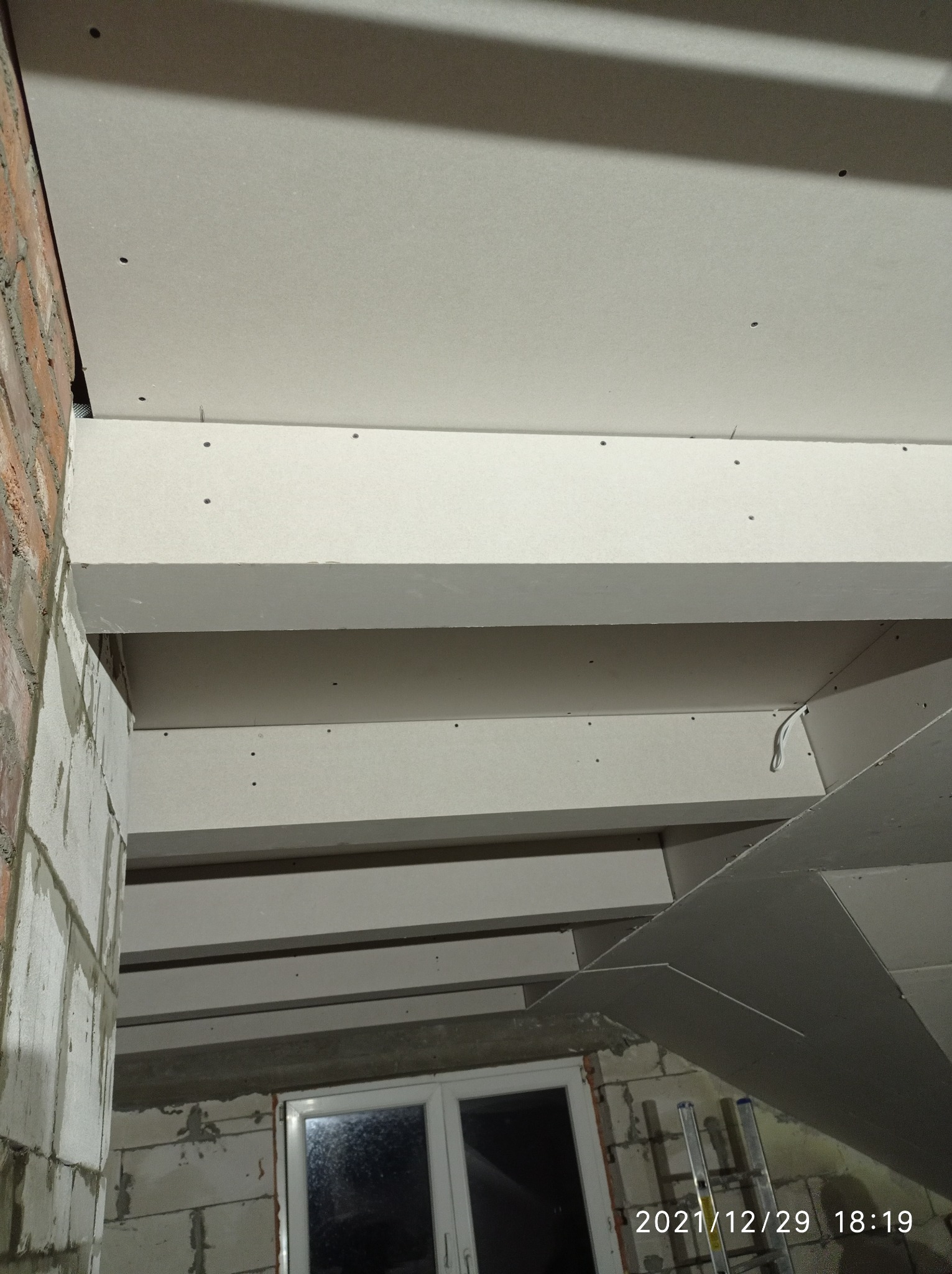 Montaż sufitu podwieszanego z płyt kartonowo-gipsowych, widoczne profile nośne, częściowo wykończona ściana z cegły i okno w tle.