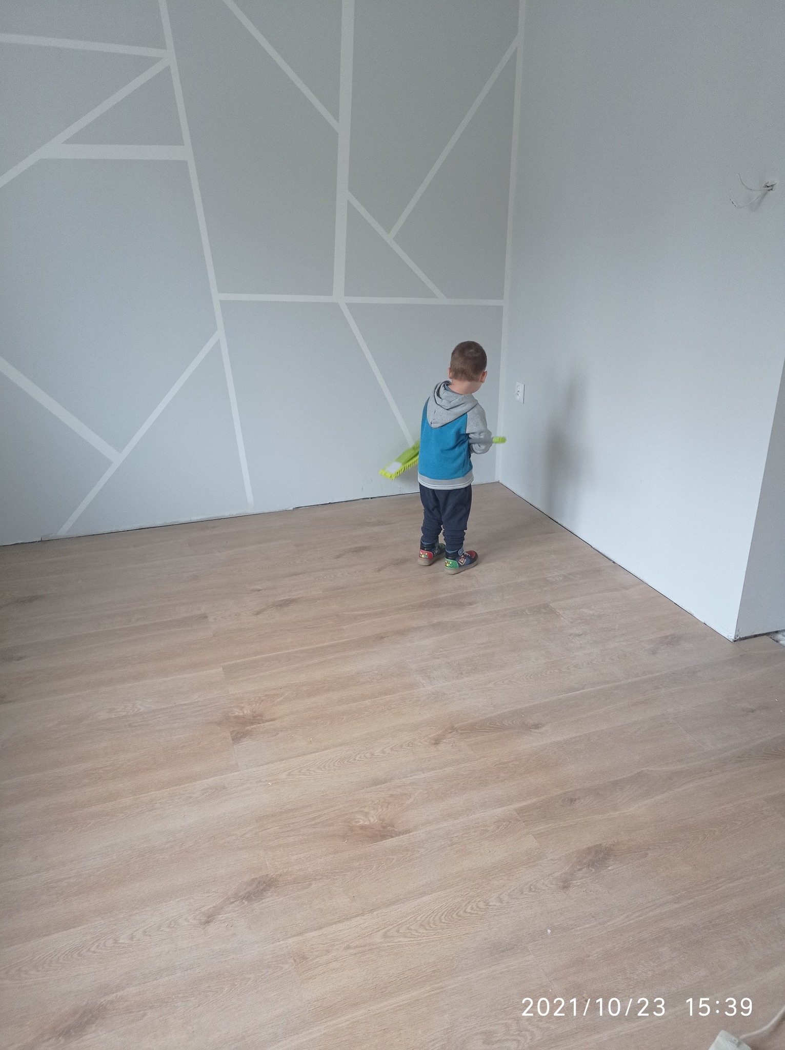 Pokój po remoncie z nową podłogą z paneli i pomalowaną ścianą z geometrycznym wzorem, mały chłopiec z zabawkami w tle.
