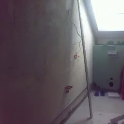 Ściana w trakcie prac przygotowawczych: widoczne przewody elektryczne, rury instalacji wodnej. W tle okno dachowe i fragment bojlera. Listwa przypodłogowa częściowo odspojona.