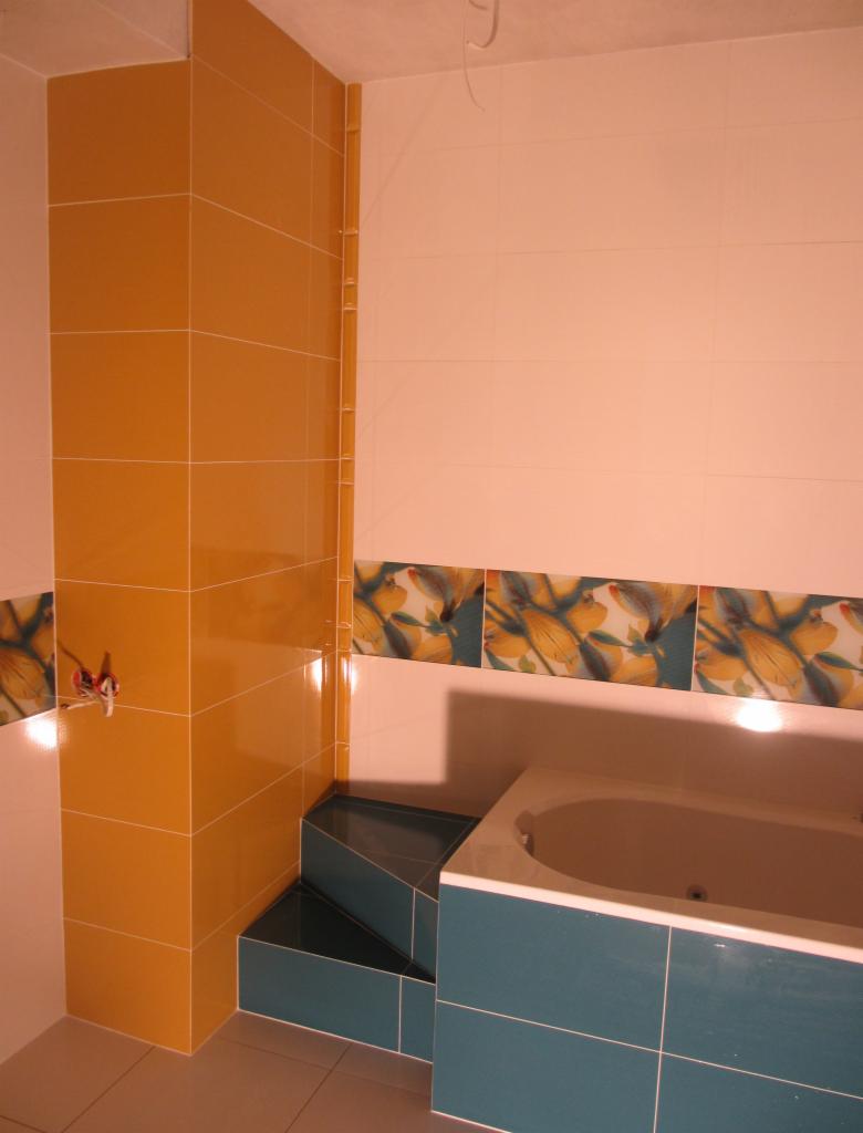 Wykończona łazienka z wanną, obłożona glazurą w odcieniach żółci i turkusu, z dekoracyjnym pasem kwiatowym. Widoczne schodki obłożone płytkami.