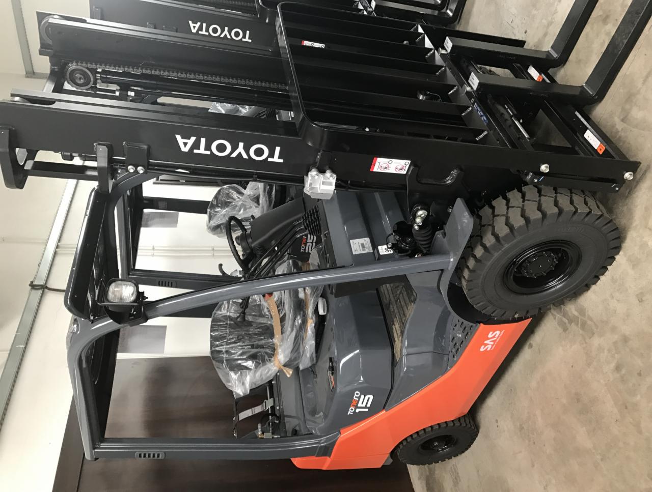 Nowy wózek widłowy Toyota 1.5, szaro-pomarańczowy, z czarnym masztem i widłami, zaparkowany w magazynie, widoczne folie ochronne na siedzeniu i kierownicy.