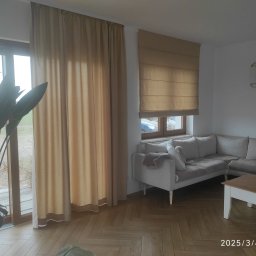 LULU JUSTYNA KIEDROWSKA - Beżowe zasłony i roleta rzymska w jasnym salonie z parkietem w jodełkę, sofa narożna i roślina doniczkowa. Widoczne detale wykończenia okien.