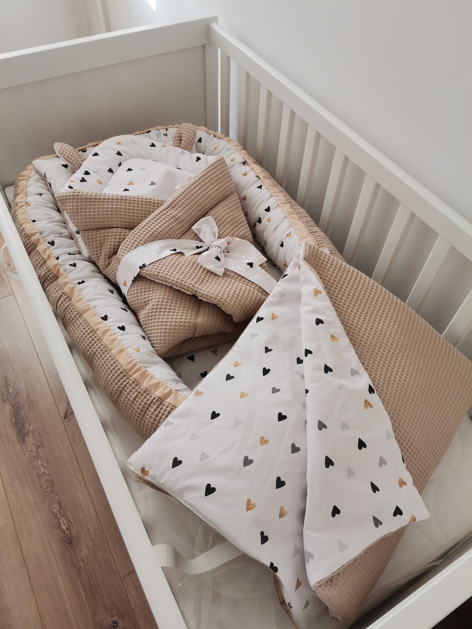 Beżowy kokon niemowlęcy z białymi elementami w serduszka, w białym łóżeczku. Waflowa faktura materiału. Zestaw pościeli dziecięcej w neutralnych barwach.