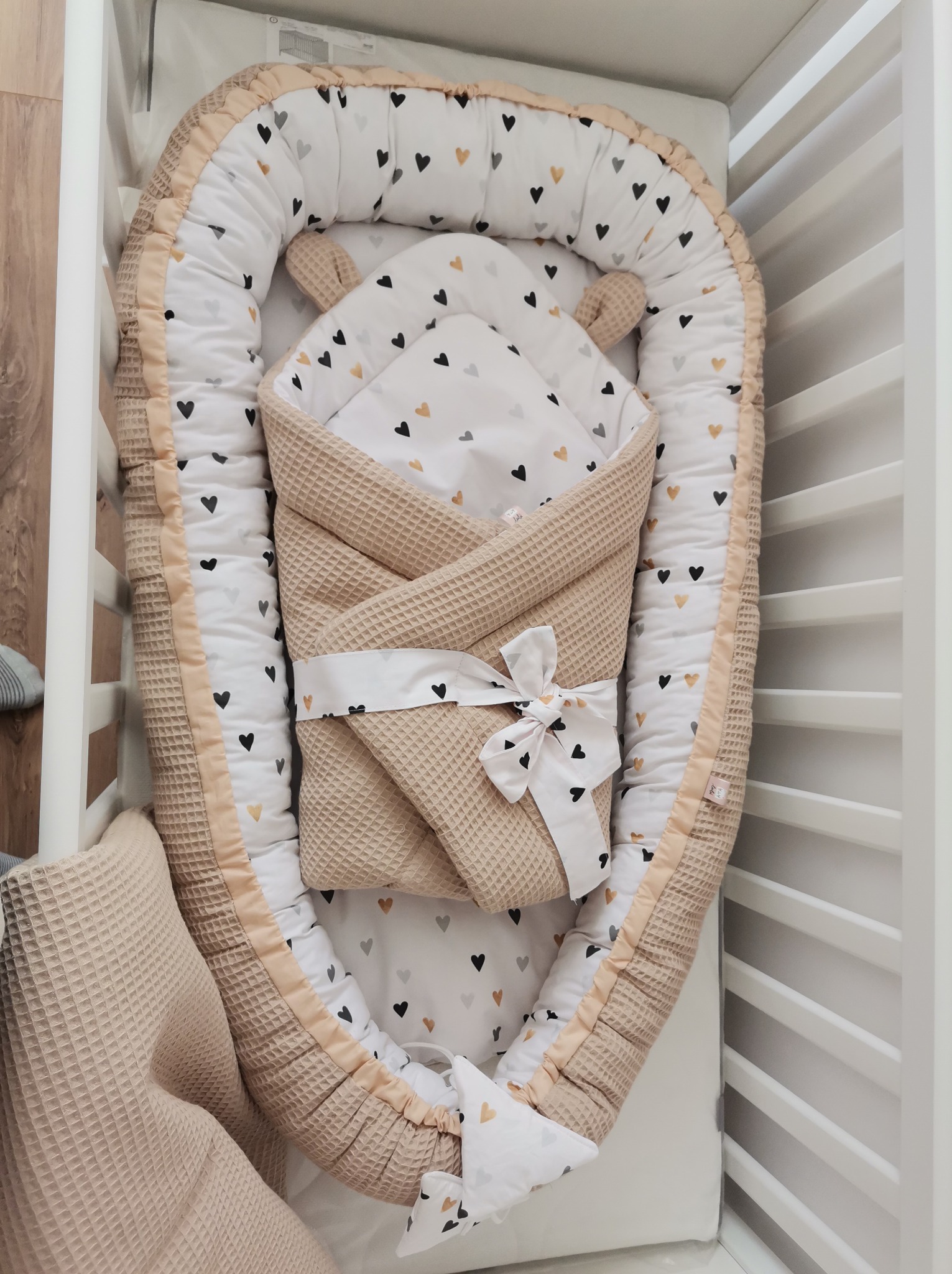 Beżowy kokon niemowlęcy z serduszkami w łóżeczku, otulony waflowym kocykiem z kokardą. Wnętrze łóżeczka wyściełane wzorem w serca. Ujęcie z góry, delikatne światło.
