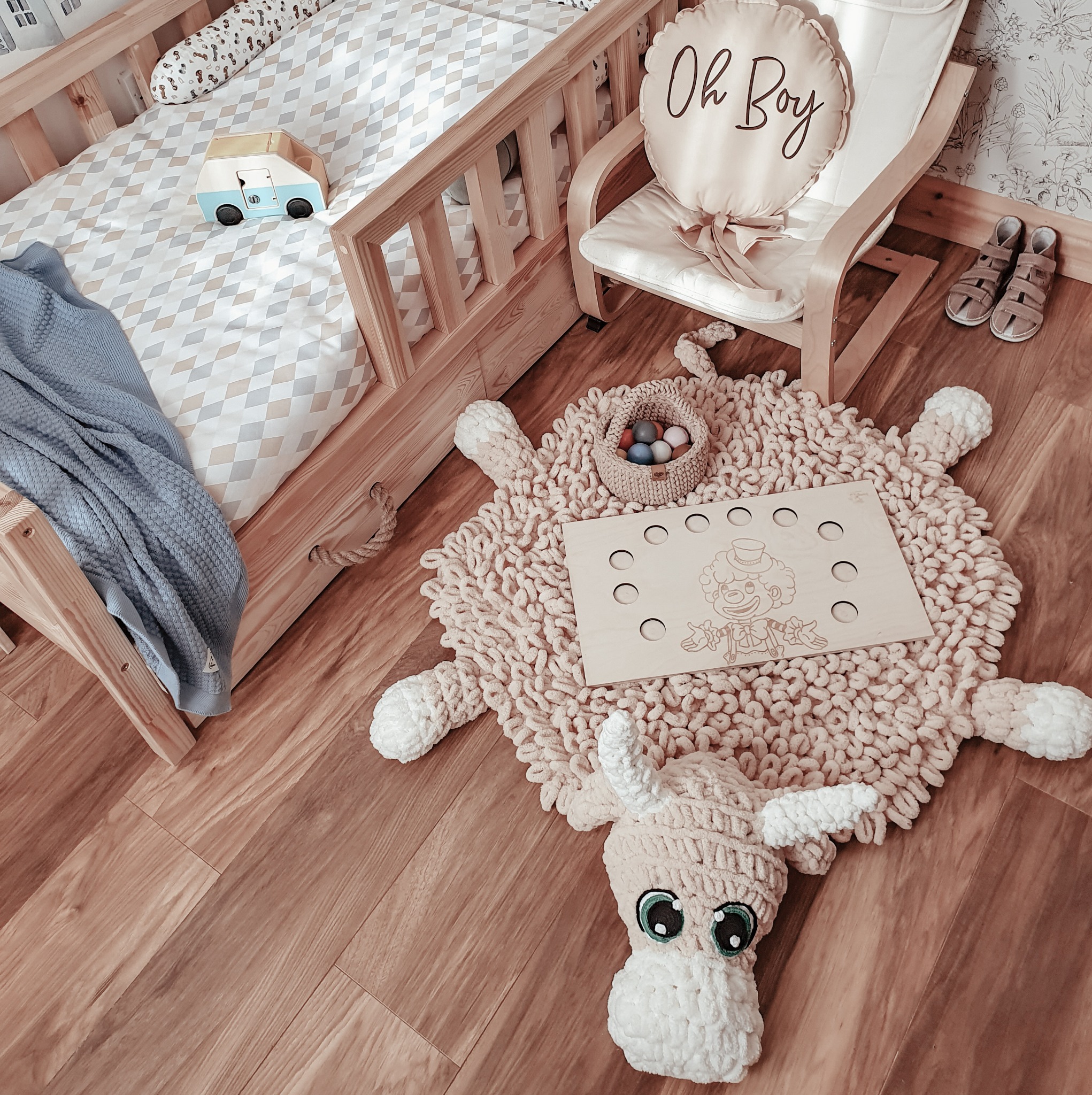 Pokój dziecięcy z drewnianym łóżeczkiem, dywanem w kształcie owcy i krzesłem z poduszką 'Oh Boy'. Widoczna drewniana podłoga i dziecięce sandałki.