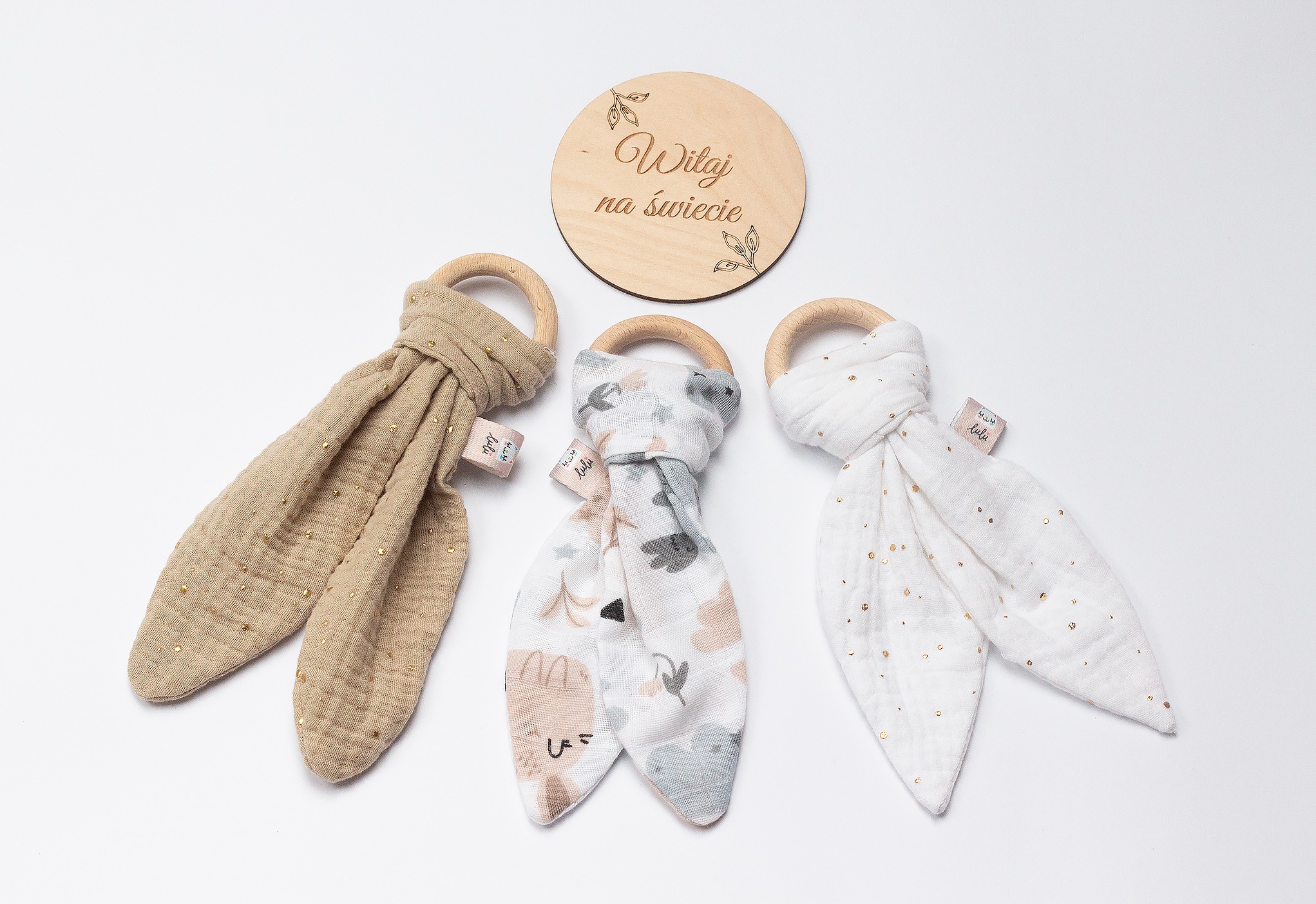 Trzy gryzaki z bawełny muślinowej z drewnianymi pierścieniami i tabliczka z napisem „Witaj na świecie” na białym tle. Różne wzory materiałów.