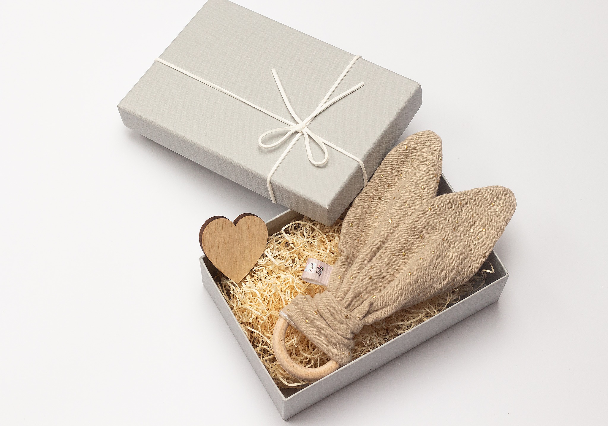 Beżowy gryzak z uszami królika w szarym pudełku prezentowym z drewnianym sercem, wypełnionym siankiem. Ujęcie z góry, na białym tle, z widoczną fakturą materiału.
