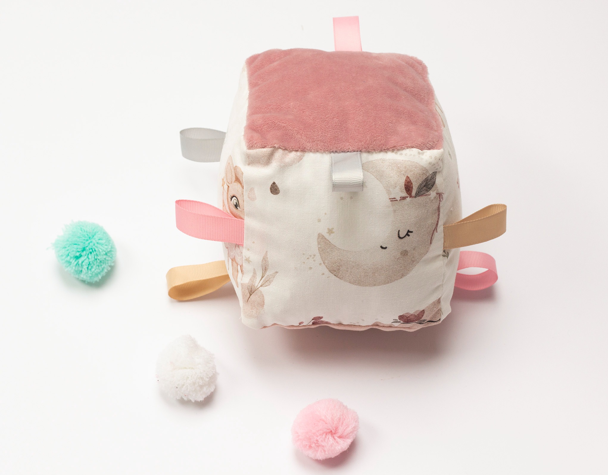 Miękki, sensoryczny sześcian dla niemowląt z różowym welurem i wstążkami, leżący na białym tle z kolorowymi pomponami. Idealny prezent dla dziecka.