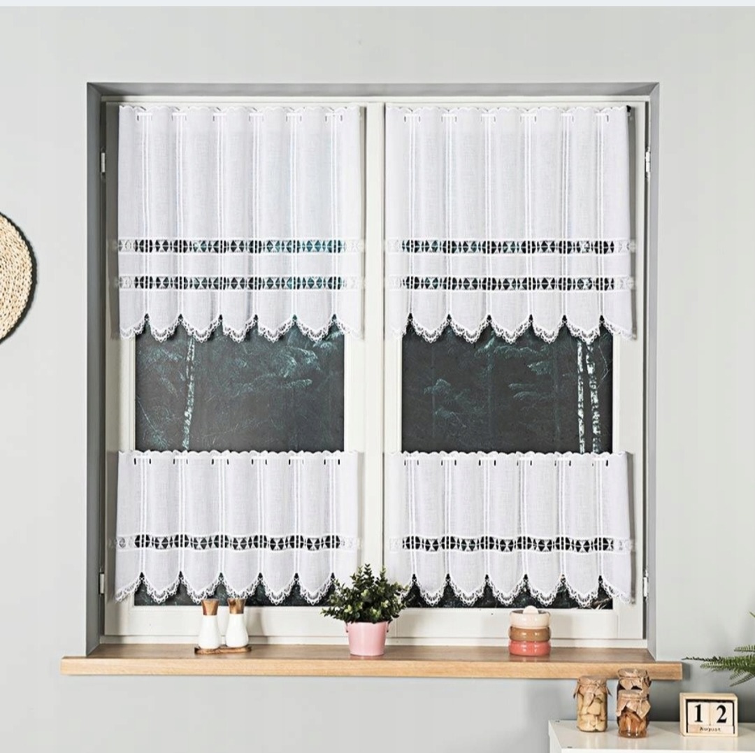 Białe, koronkowe zazdrostki w oknie, z dekoracyjnym wykończeniem. Na parapecie ozdoby, kwiat doniczkowy i kalendarz. Widok na las za oknem.