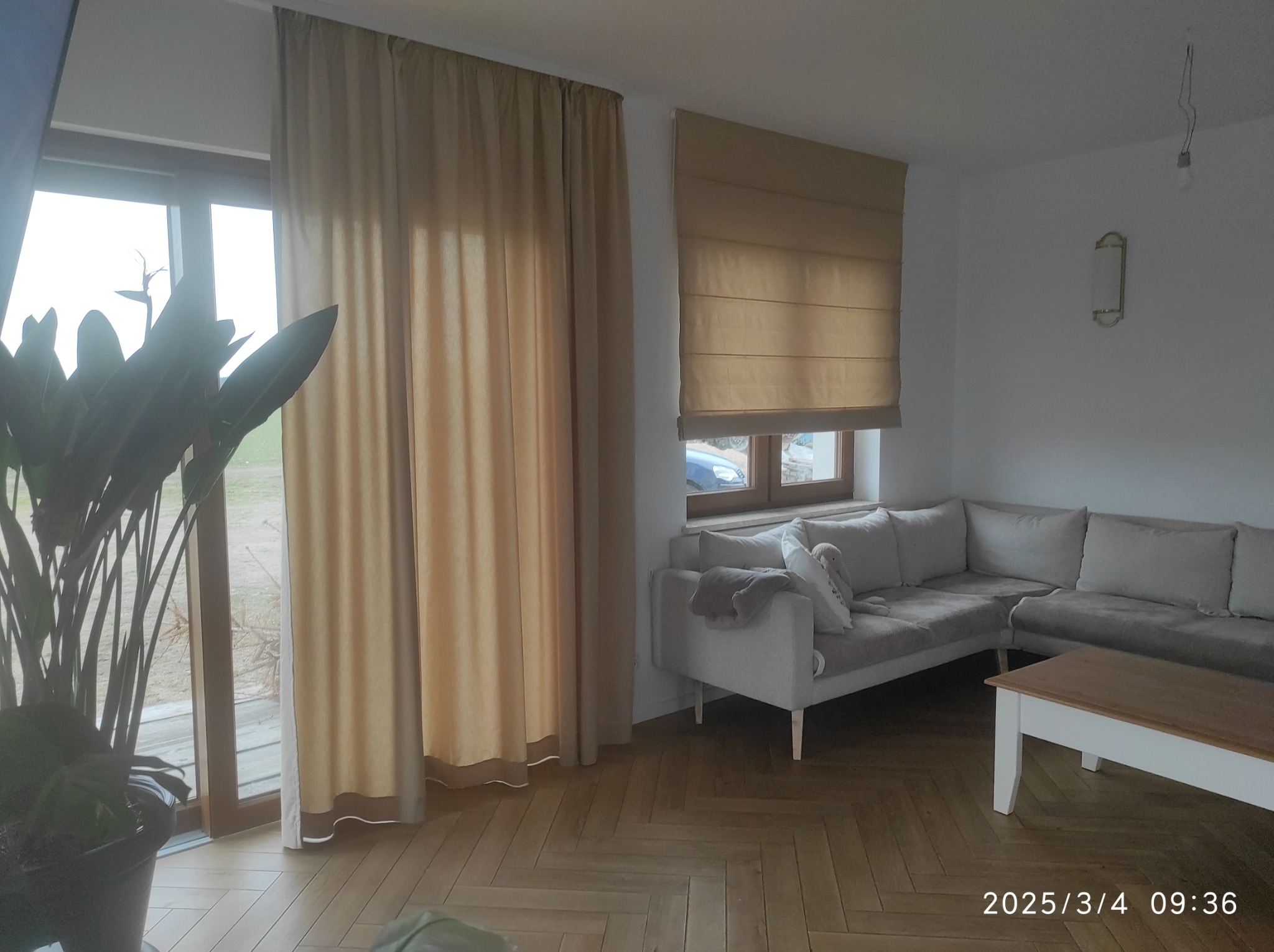 Beżowe zasłony i roleta rzymska w jasnym salonie z parkietem w jodełkę, sofa narożna i roślina doniczkowa. Widoczne detale wykończenia okien.