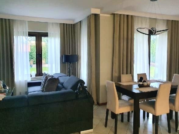 Elegancki salon z szarymi zasłonami i firanami, ciemnym narożnikiem oraz stołem jadalnym. Jasne krzesła i nowoczesna lampa dodają wnętrzu lekkości.