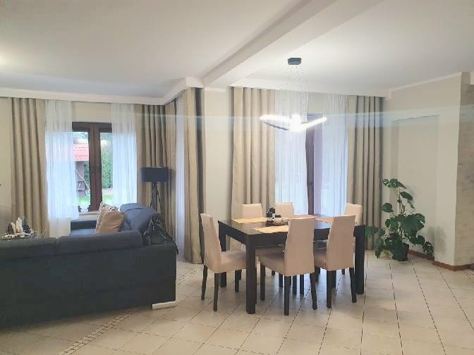 Eleganckie zasłony w salonie z dużym oknem, beżowe i białe, tworzą przytulną atmosferę. Widoczny stół z krzesłami i nowoczesna lampa.