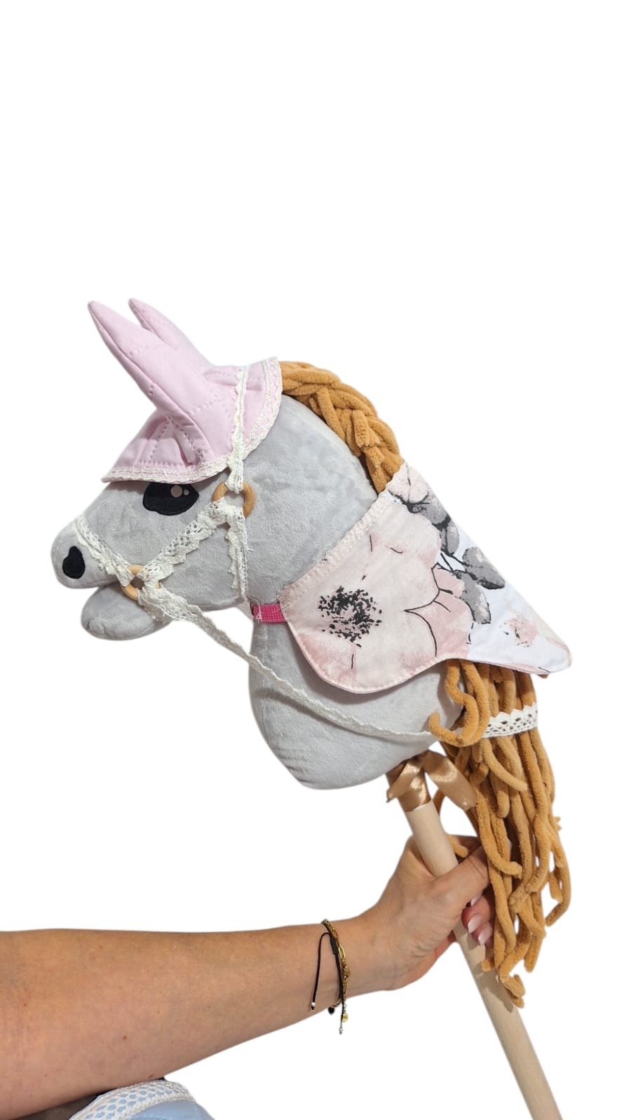 Ręcznie szyty, pluszowy koń na kiju w pastelowych barwach, z grzywą z włóczki i dekoracyjnym czaprakiem w kwiaty, trzymany przez osobę.