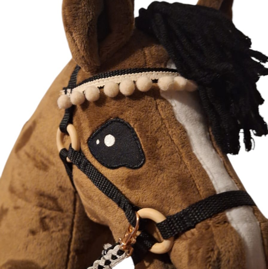 Pluszowy koń na kiju z czarną grzywą i ozdobnym kantarem z pomponami, zbliżenie na głowę z widocznymi detalami szycia i materiałów.