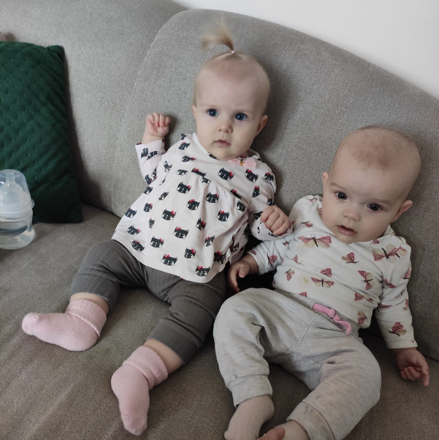 Dwie urocze dziewczynki w uszytych w Chojnicach ubrankach z motywami kotków i motyli siedzą na szarej kanapie, obok butelka. Pastelowe kolory, miękkie światło.