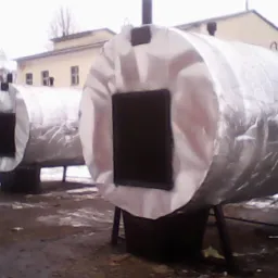 Dwa cylindryczne zbiorniki z izolacją termiczną i czarnymi otworami na metalowych nogach, na zewnątrz, w tle budynek i drzewa bez liści.