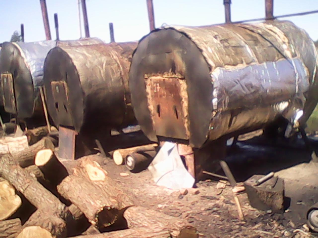 Rzędy metalowych beczek na paleniskach, częściowo owiniętych folią, z przygotowanym drewnem do palenia. Widoczne narzędzia. Produkcja na zewnątrz.