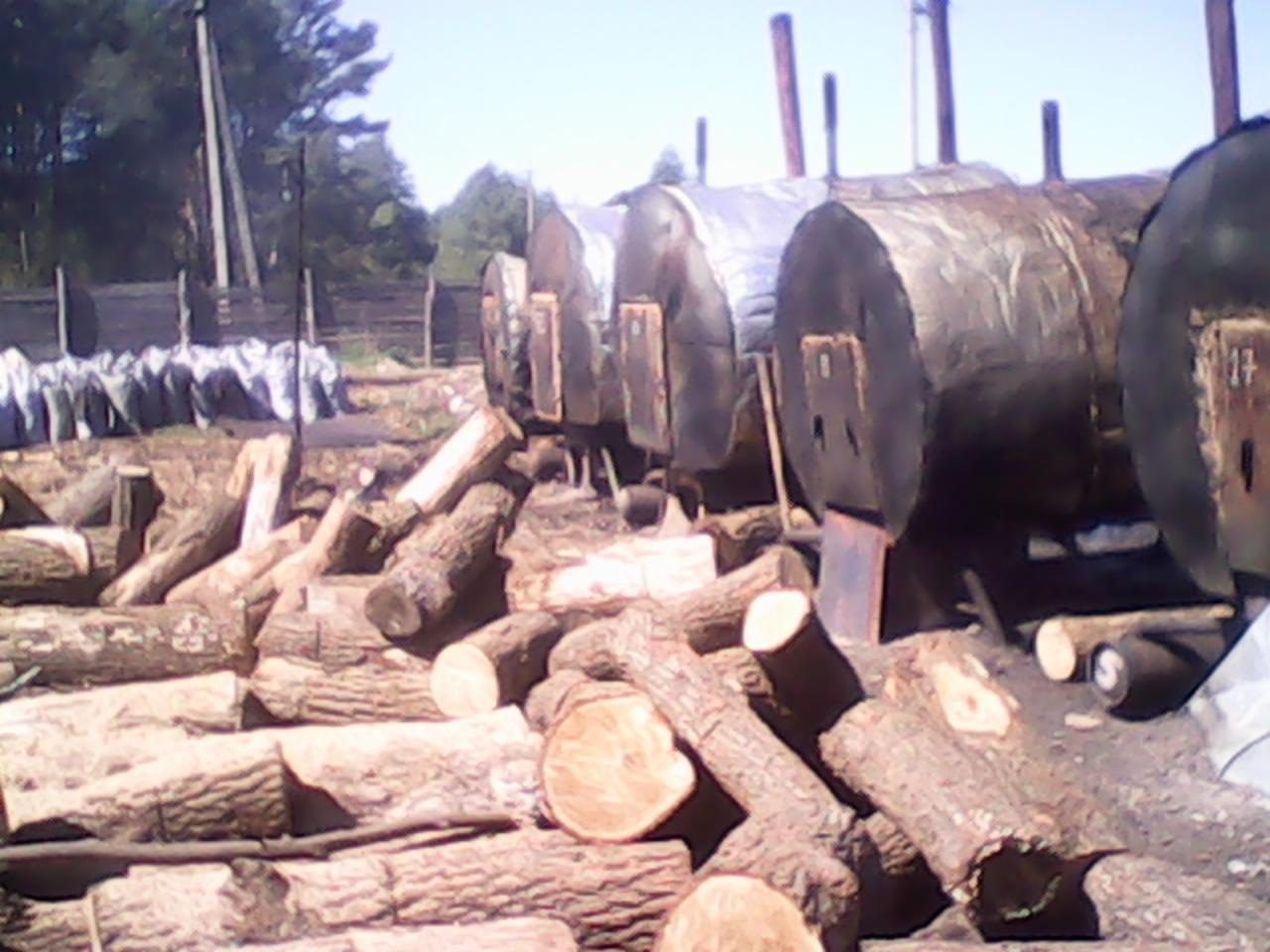 Rząd metalowych zbiorników z kominami i stosem drewna opałowego na zewnątrz, prawdopodobnie w trakcie procesu produkcji lub przetwarzania.