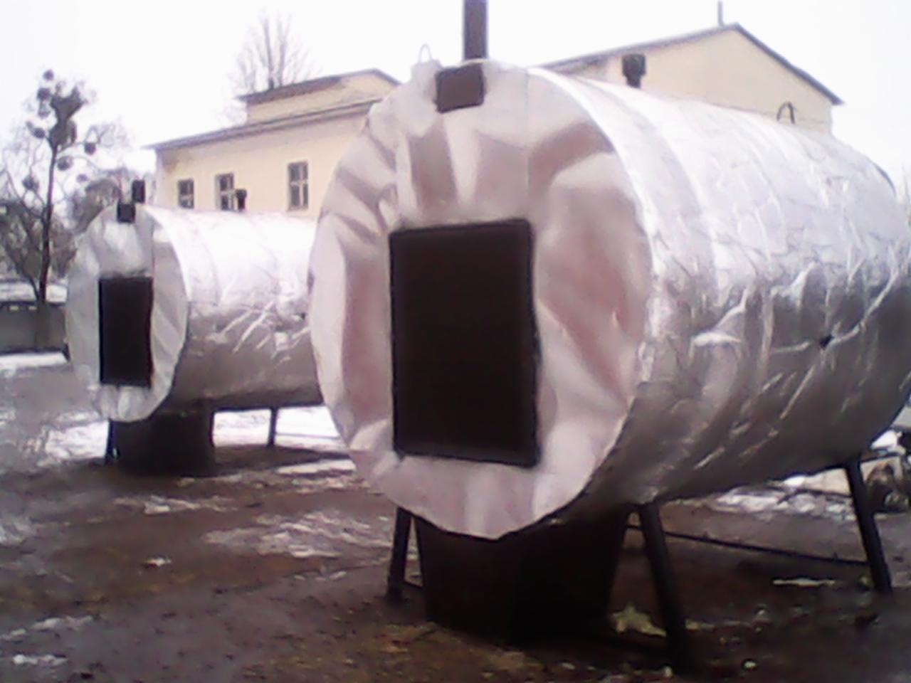 Dwa cylindryczne zbiorniki z izolacją termiczną i czarnymi otworami na metalowych nogach, na zewnątrz, w tle budynek i drzewa bez liści.