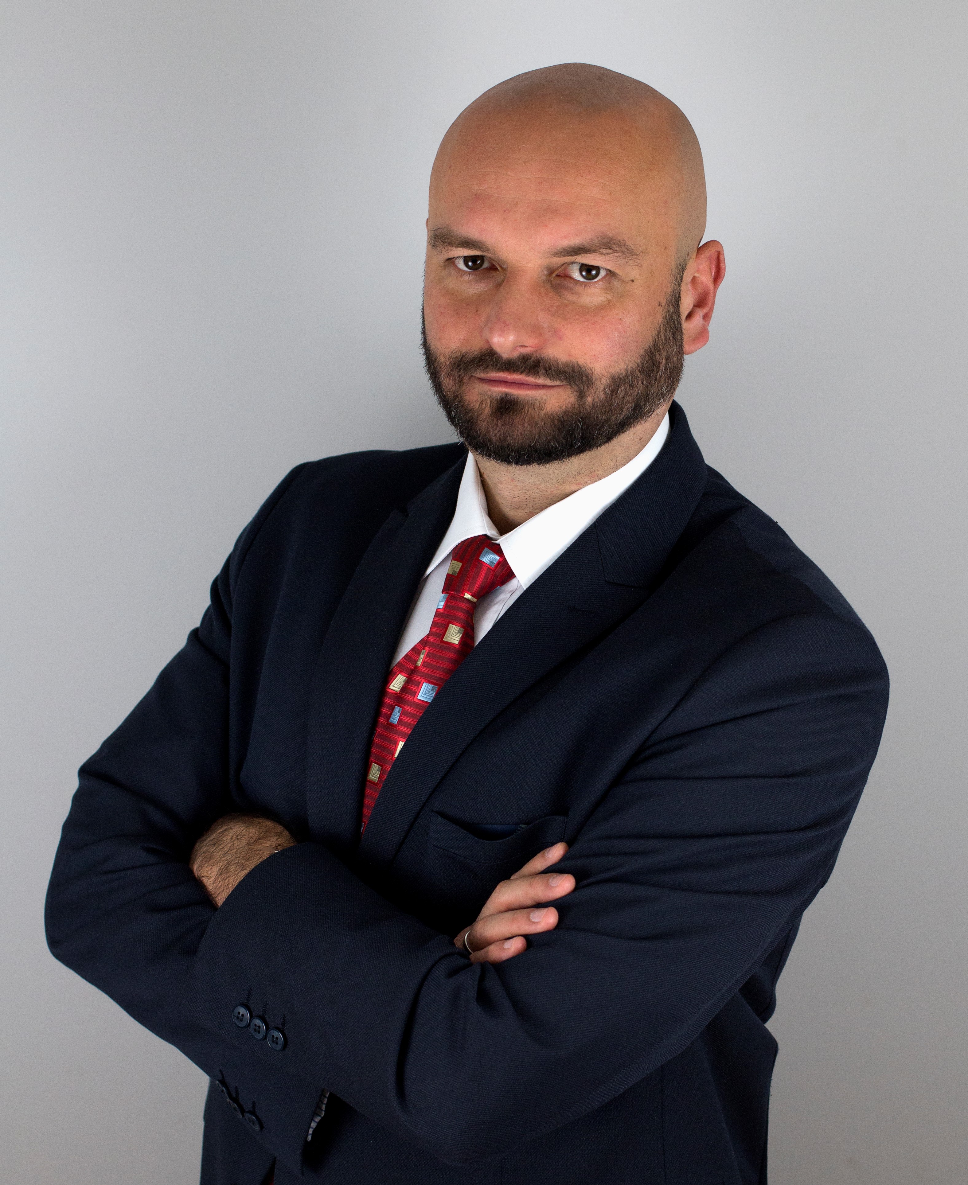 Portret mężczyzny w garniturze z założonymi rękami na tle szarej ściany w Warszawie. Mężczyzna ma brodę i krawat w geometryczne wzory.