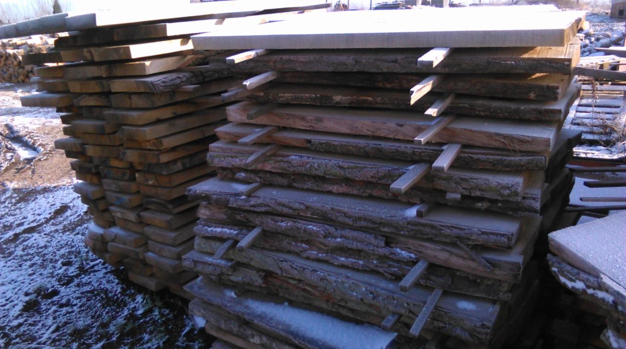 Stos desek z korą, przełożonych listewkami, na zewnątrz lekko przyprószone śniegiem. Widoczne ślady obróbki drewna, jasne oświetlenie dzienne.