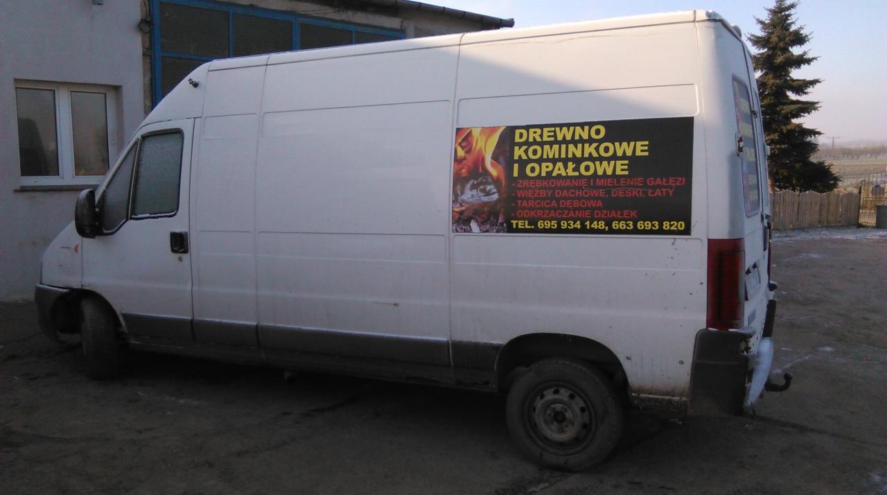 Biały bus z reklamą sprzedaży drewna kominkowego i opału, z informacjami o zrębkowaniu gałęzi, więźbach dachowych i tarcicy dębowej. Widoczny numer telefonu.