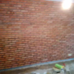 Ściana wykończona czerwoną cegłą dekoracyjną, widoczne narzędzia i akcesoria malarskie, drabina. Nierównomierne oświetlenie, surowy stan podłoża, wnętrze w trakcie prac wykończeniowych.