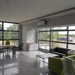 Wnętrze biura w trakcie wykańczania, widoczny zamontowany klimatyzator ścienny, instalacje elektryczne wystające z sufitu, duże okna z widokiem na zewnątrz.