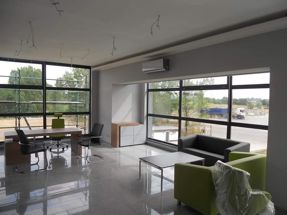 Wnętrze biura w trakcie wykańczania, widoczny zamontowany klimatyzator ścienny, instalacje elektryczne wystające z sufitu, duże okna z widokiem na zewnątrz.