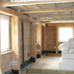 Wnętrze drewnianego domu w trakcie budowy lub remontu, widoczne drewniane ramy okienne, surowy strop, ściany z desek, izolacja fundament&oacute;w i stos białych bloczk&oacute;w betonowych.