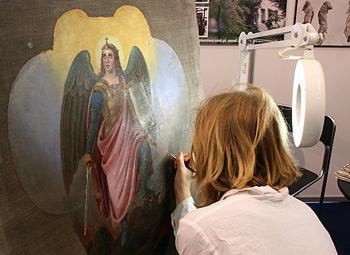 Artysta konserwator z bliska, podczas retuszu malowidła religijnego przedstawiającego postać anioła na płótnie, używa cienkiego pędzla w oświetlonym stanowisku pracy.