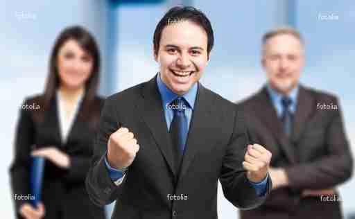 Uśmiechnięty mężczyzna w garniturze, z zaciśniętymi pięściami, wyrażający radość, z dwoma rozmazanymi współpracownikami w tle.