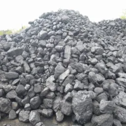 Kupa węgla kostki o nieregularnych kształtach, widoczna siatka ogrodzeniowa w tle, pochmurny dzień.