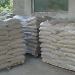 Stosy worków z pelletem na paletach, zabezpieczone folią, ustawione w magazynie. Widoczne surowe ściany i okno w tle.