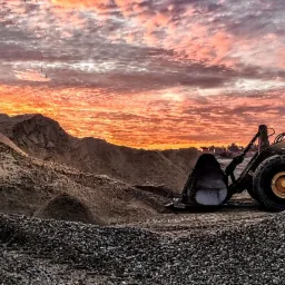 Żółta ładowarka Volvo na tle hałd żwiru i piasku podczas zachodu słońca, widoczna taśma podajnika