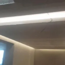 Podwieszany sufit z oświetleniem LED w kształcie prostokąta, widok z dołu na szarą powierzchnię sufitu i jasne, białe światło. W tle okno.