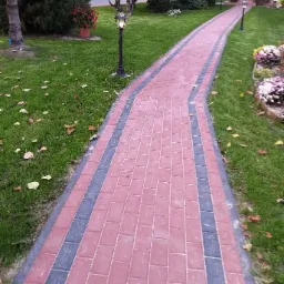 Wąska ścieżka z czerwonej kostki brukowej z szarymi obrzeżami, wijąca się przez zielony trawnik z opadłymi liśćmi, oświetlona lampami ogrodowymi.