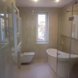 Nowoczesna łazienka w jasnych odcieniach z wolnostojącą wanną, podwieszaną toaletą i grzejnikiem drabinkowym na ścianie, widok zza szklanej kabiny prysznicowej.