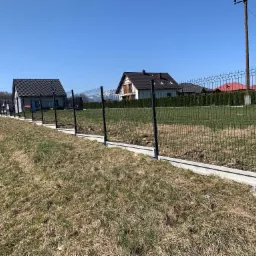 Nowo zainstalowane, czarne ogrodzenie z paneli na betonowej podmurówce w Milówce, z widokiem na domy i błękitne niebo w tle.