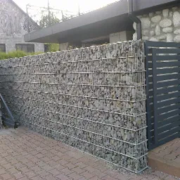 Gabionowe ogrodzenie z szarego kamienia i metalowa brama, tworzące nowoczesny design. Widoczne elementy konstrukcyjne i faktura kamienia.